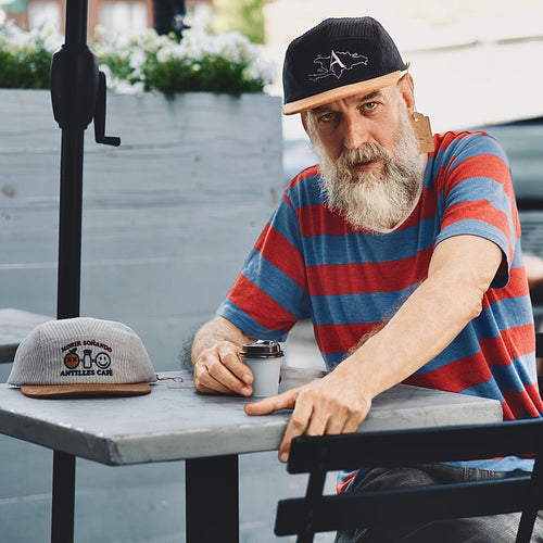 Man with a beard wearing a striped shirt and cap sitting at an outdoor table with a cap and cup.