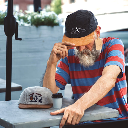Man sitting at a table with hats and a coffee cup, wearing a striped shirt and cap.