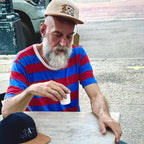 Man in a striped shirt and cap sitting outdoors, holding a cup.