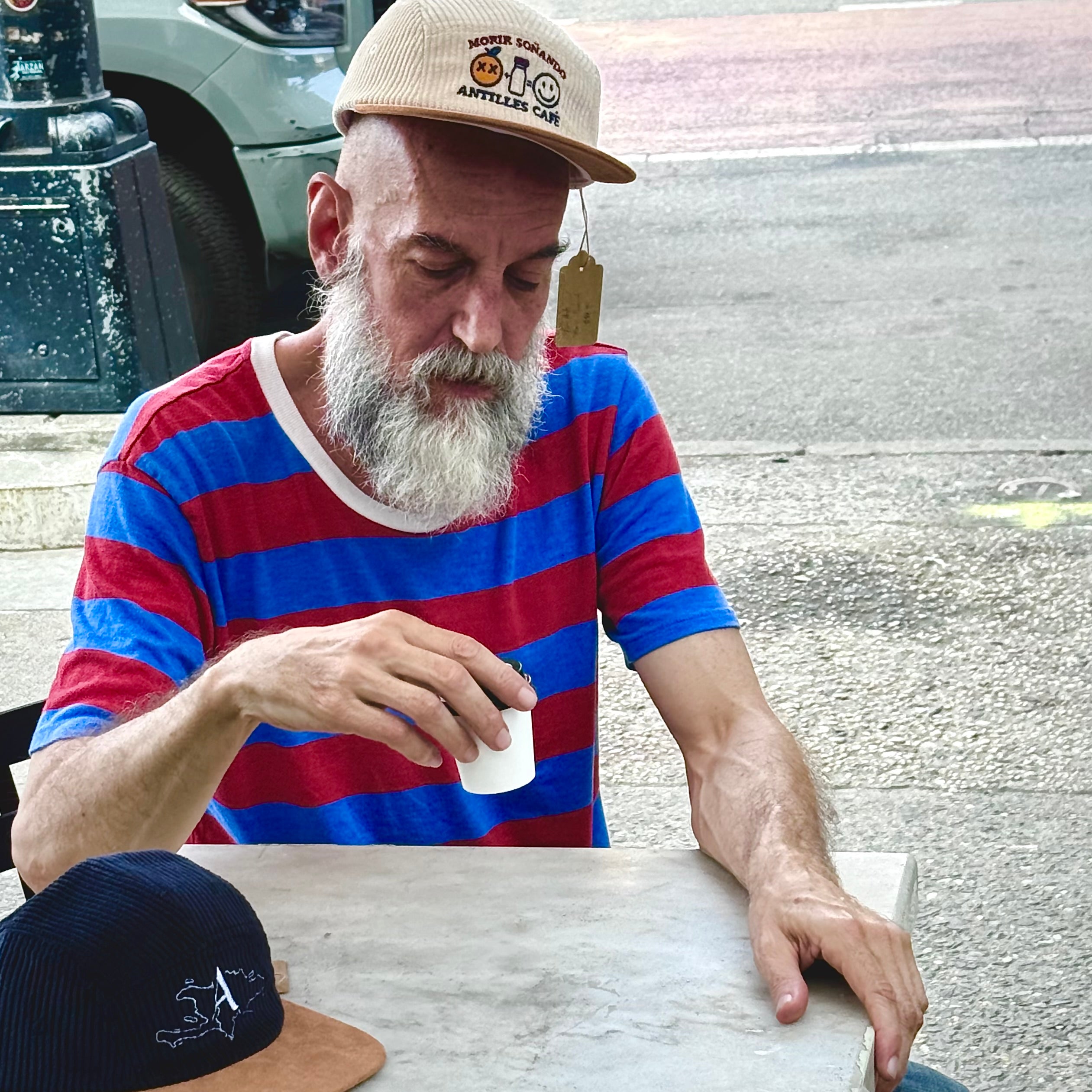 Man in a striped shirt and cap sitting outdoors, holding a cup.