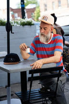 Man sitting at an outdoor table with a cup, wearing a striped shirt and cap.