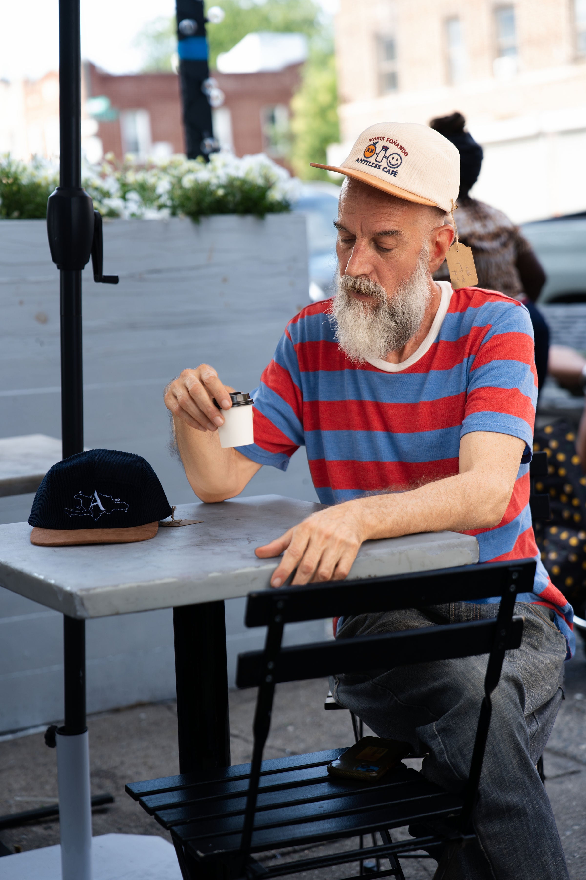 Man sitting at an outdoor table with a cup, wearing a striped shirt and cap.